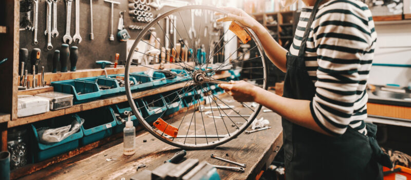 Cute Caucasian female worker holding and repairing bicycle wheel while standing in bicycle workshop.