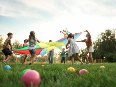 Group of children and teachers playing with rainbow playground parachute on green grass, low angle view. Summer camp activity